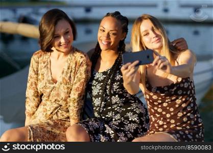 Group of delighted multiracial female friends in dresses taking selfie on smartphone while sitting in port on sunny summer day. Young diverse women taking self portrait
