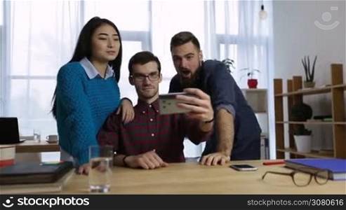 Group of diverse cheerful coworkers taking self portrait and making funny gestures in open space office. Smiling hipster colleagues in casual outfits making selfie on smartphone at workplace. Dolly.