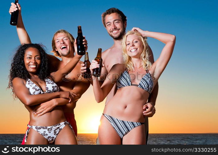 Group of four friends - men and women - standing with drinks on the beach against the setting sun over the ocean