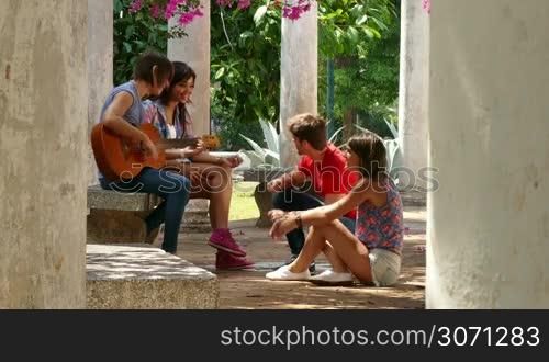 Group of four friends, students free time, youth in summer, young people outdoor, men and women having fun in city park, leisure, relax, lifestyle. Man playing music with guitar and girls singing tune