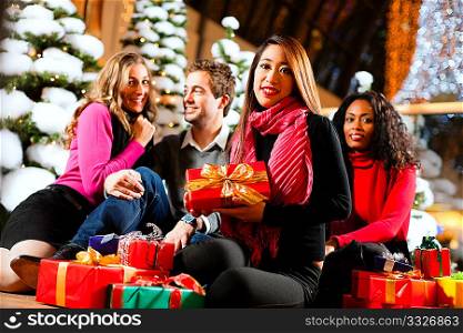 Group of four people - diversity - sitting amid artificial snow covered fir trees and lights with Christmas presents in a shopping mall