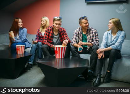 Group of friends eating popcorn and waiting for showtime in cinema hall. Male and female youth sitting on sofa in movie theater