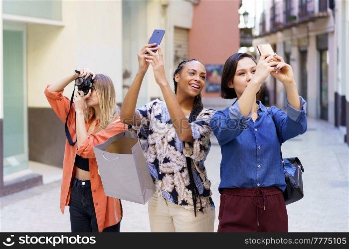 Group of glad diverse female friends taking pictures of street with smartphones and photo camera while standing on sidewalk near buildings. Content multiracial friends taking photos of street