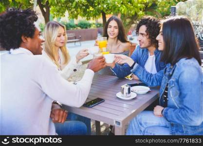 Group of happy diverse friends clinking cups and glasses while gathering at table in outdoors cafe and celebrating meeting in summer. Company of multiethnic friends celebrating reunion