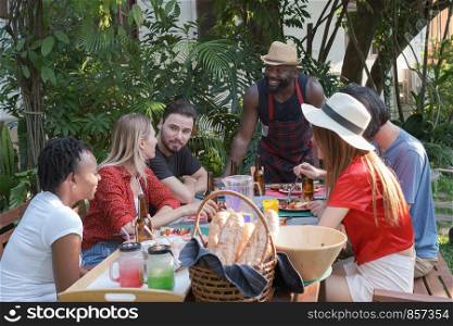 Group of happy friends eating and drinking beers at barbecue dinner camping in nature and having meal together outdoor as summer lifestyle, food and friendship concept