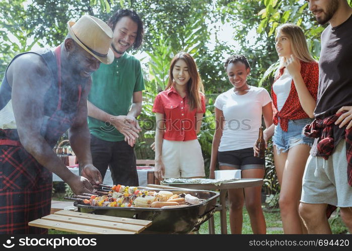 Group of happy friends standing eating and drinking beers at barbecue dinner camping in nature and having meal together outdoor as summer lifestyle, food and friendship concept