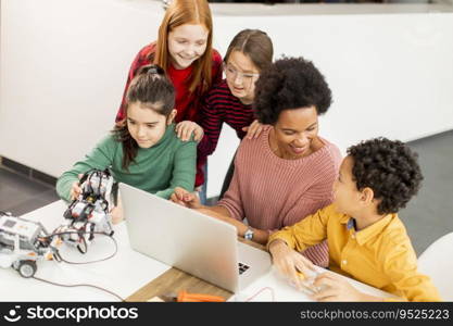 Group of happy kids with their African American female science teacher with laptop programming electric toys and robots at robotics classroom