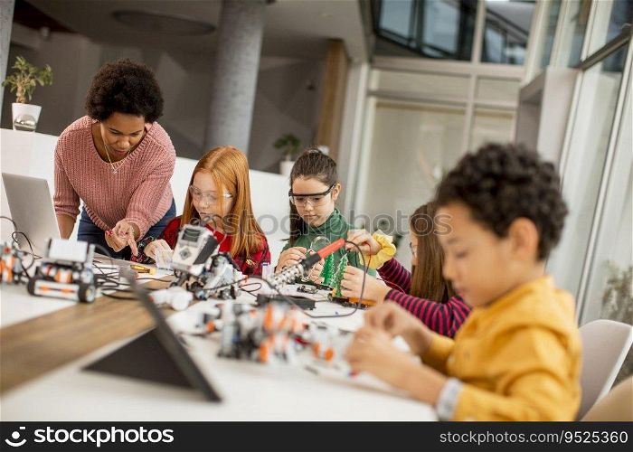 Group of happy kids with their African American female science teacher with laptop programming electric toys and robots at robotics classroom
