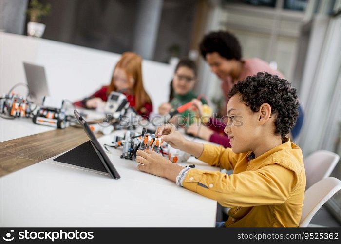 Group of happy kids with their African American female science teacher with laptop programming electric toys and robots at robotics classroom