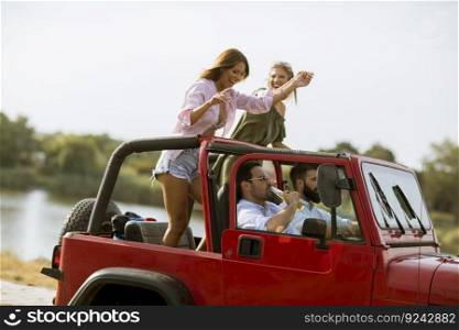 Group of happy young friends having fun in convertible car during summer vacation by river