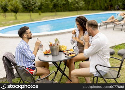 Group of happy young people cheering with drinks and eating fruits by the pool in the garden