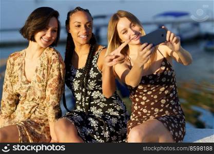 Group of joyful female friends in dresses taking self portrait on smartphone while sitting together on pier on summer day. Positive diverse women taking selfie near river