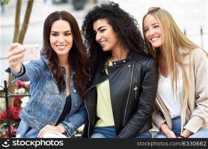 Group of multi-ethnic young women taking a selfie photograph together outdoors. Blonde, brunette and mixed females wearing casual clothes in urban background.