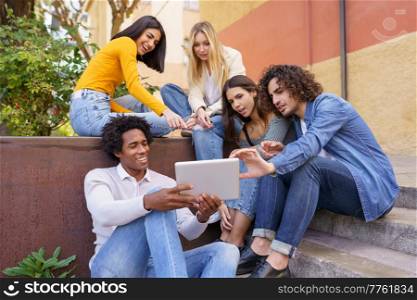Group of multiethnic best friends gathering in city and watching funny video on tablet while spending time together. Company of diverse friends watching video on tablet