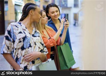 Group of multiethnic female friends taking picture of showcase on smartphone while standing together near boutique on street during shopping. Multiracial women taking photo of showcase in city