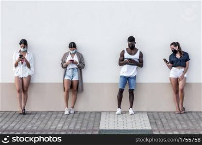 Group of multiethnic friends in masks standing near urban building and surfing Internet on mobile phones on Lanzarote. Diverse friends browsing smartphones on street