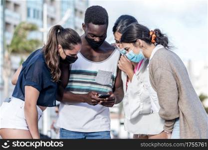 Group of multiethnic friends in masks standing on street and watching funny video on mobile phone while enjoying weekend together on Lanzarote. Company of diverse friends using smartphone together in city