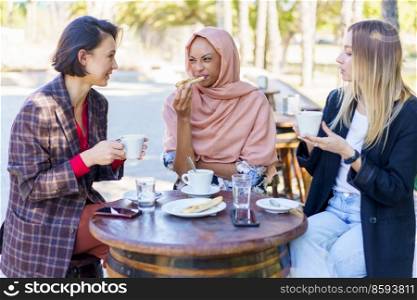 Group of multiracial female friends having lunch together at table with cups of coffee on terrace of outdoor cafe in city. Diverse women having lunch in city
