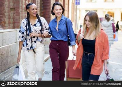 Group of optimistic multiethnic female friends with shopping bags strolling together on paved street near building after shopping in city. Content multiracial women with shopping bags