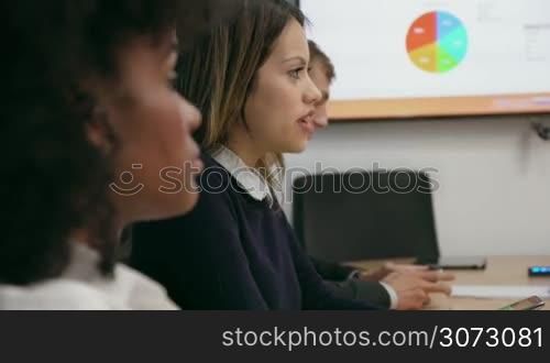 Group of people during business meeting in corporate conference room. One caucasian and one african american woman talk with colleagues with charts on TV in background. Closeup shot
