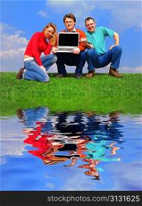 Group of people with notebook sitting on grass against sky with reflection on water
