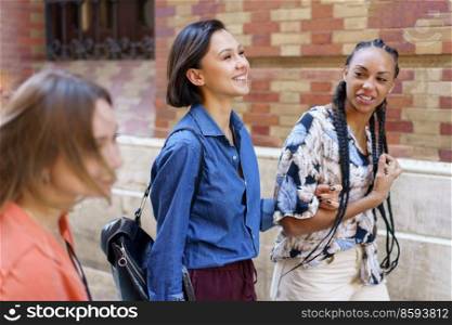 Group of positive multiracial female friends in colorful wear strolling together on street near brick building in city on summer day. Cheerful diverse women walking on street