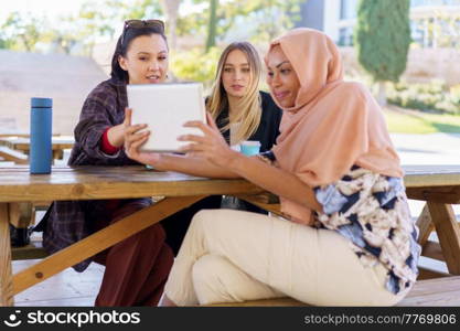 Group of positive young multiethnic women smiling and drinking coffee, to go while watching funny video on tablet, during break in outdoor cafe in park. Cheerful diverse women sharing tablet during coffee break in cafe