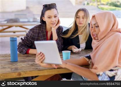 Group of positive young multiethnic women smiling and drinking coffee, to go while watching funny video on tablet, during break in outdoor cafe in park. Cheerful diverse women sharing tablet during coffee break in cafe