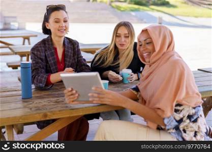 Group of positive young multiethnic women smiling and drinking coffee, to go while watching funny video on tablet, during break in outdoor cafe in park. Cheerful diverse women sharing tablet during coffee break in cafe
