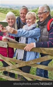 Group Of Senior Friends Hiking In Countryside Standing By Gate  And Taking Selfie On Mobile Phone                                     