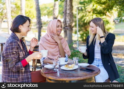 Group of smiling diverse female friends chatting while having coffee break at table on terrace of outdoor cafeteria in city. Cheerful multiracial women having coffee break