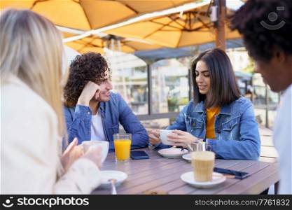 Group of smiling multiracial friends gathering around table in outdoors cafe and chatting while enjoying meeting at summer weekend. Company of diverse friendly people in summer cafe