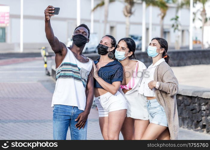 Group of smiling multiracial friends wearing masks standing in city and taking selfie on smartphone while spending time together in summer on Lanzarote. Company of diverse friends in masks taking selfie on street