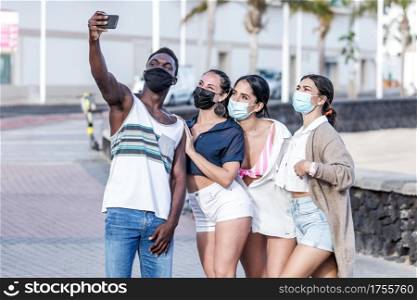 Group of smiling multiracial friends wearing masks standing in city and taking selfie on smartphone while spending time together in summer on Lanzarote. Company of diverse friends in masks taking selfie on street
