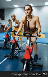 Group of sportive women doing exercise on stationary bikes in gym, front view. People on fitness workout in sport club, athletic girls in sportswear on training indoors. Women on stationary bikes in gym, front view