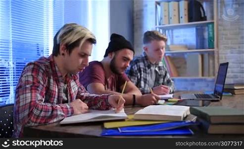 Group of students studying together in class and taking lecture notes to notebooks with pen and pencil. Handsome teenagers writing lecture on notepads while learning at college.