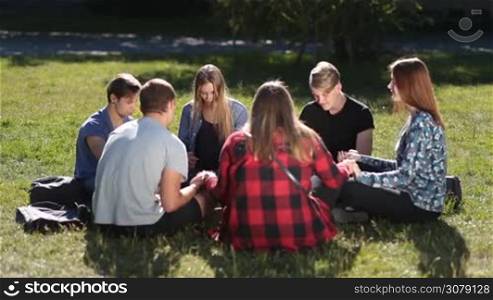 Group of teenage college christian friends sitting in circle on green grass and praying. Cheerful students holding hands and meditating while sitting in circle on park lawn on university campus.