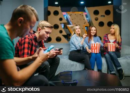 Group of teenagers relax on the couch and waiting for the film in cinema hall. Male and female youth sitting on sofa in movie theater, popcorn on the table