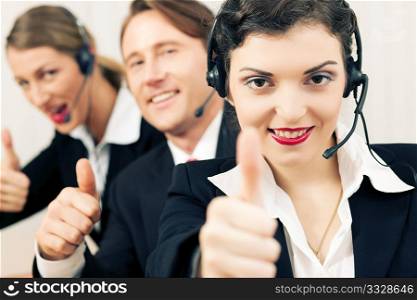 Group of three customer care representatives in a call center with headphones, all showing thumbs up