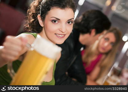 Group of three friends in a bar drinking beer - selective focus on beautiful woman in front pointing her glass at the viewer