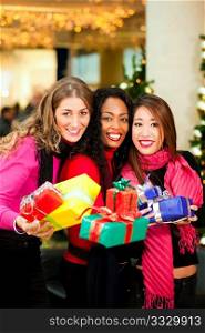 Group of three women - white, black and Asian - with Christmas presents in a shopping mall in front of a Christmas tree