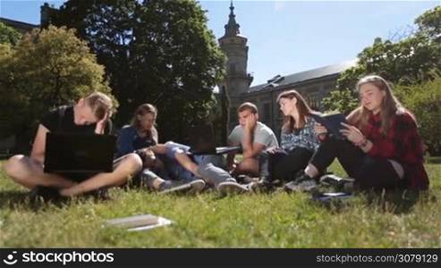 Group of tired, sleepy and exhausted college students preparing for exams on campus while sitting on green grass outside university building. Tired classmates studying hard outdoors, fighting with sleep. Dolly shot