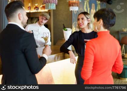 Group of well dressed friends drinking alcohol beverages and enjoying meeting while standing at wooden counter in bar with bartender. Company of elegant friends gathering in bar