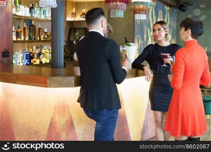 Group of well dressed friends drinking alcohol beverages and enjoying meeting while standing at wooden counter in bar with bartender. Company of elegant friends gathering in bar