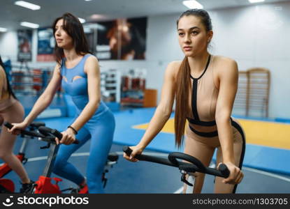 Group of women doing exercise on a stationary bikes in gym, bottom view. People on fitness workout in sport club, athletic girls in sportswear on training indoors. Women on stationary bikes in gym, bottom view