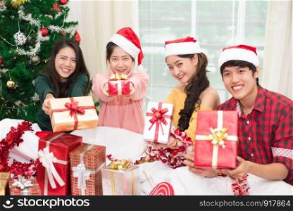 Group of young Asian people holding and giving gift box to you with Xmas tree background. Holiday and Festival concept. Christmas and New year event theme. Happy people wearing Santa hat.