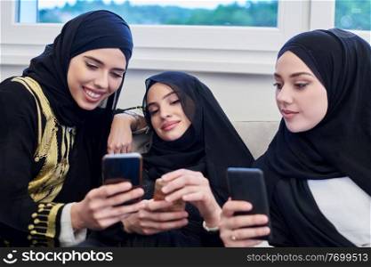group of young beautiful muslim women in fashionable dress with hijab using mobile phone while sitting on the sofa near window at home representing modern islam fashion technology and ramadan kareem concept