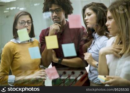 Group of young busi≠ss peop≤discussing in front of glass wall using post it notes and stickers at startup office