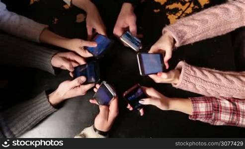 Group of young friends addicted to using mobile phone devices in cafe on black grunge table background. Upper point of view on human hands using smart phones together. Teenagers browsing social media and websites.