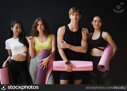 Group of young people holding yoga mats While leaning against the black wall in the gym